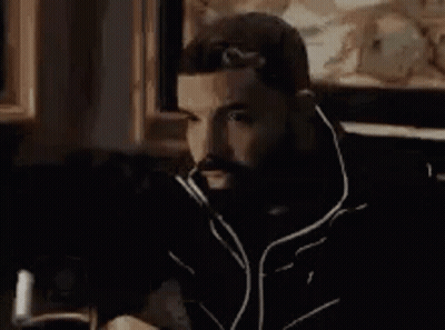 A Man With A Beard Is Sitting In A Chair In Front Of A Painting In A Room . GIF
