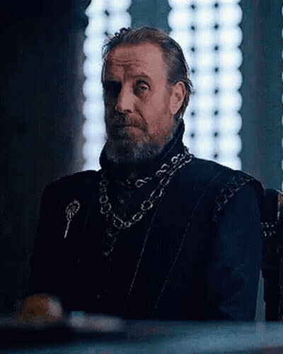 A Man With A Beard Is Sitting In A Chair In Front Of A Window Wearing A Chain Around His Neck . GIF