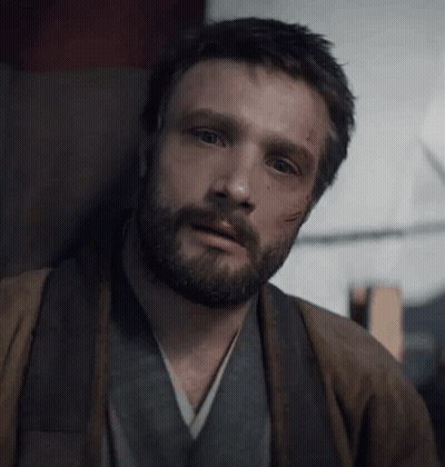 A Man With A Beard Is Sitting In A Chair Looking At The Camera . GIF