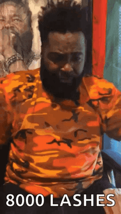 A Man With A Beard Is Sitting In A Chair With His Arms Outstretched And A Camouflage Shirt On . GIF