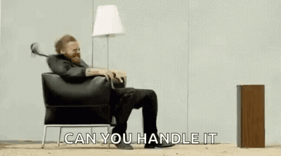 A Man With A Beard Is Sitting In A Chair With His Legs Crossed And A Lamp . GIF