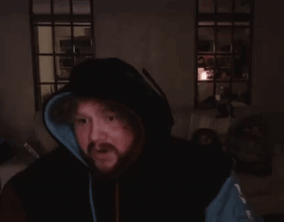 A Man With A Beard Is Sitting In A Dark Room With His Hands Folded . GIF