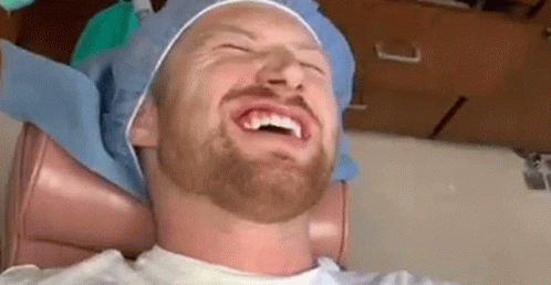 A Man With A Beard Is Sitting In A Dental Chair Laughing While A Dentist Examines His Teeth . GIF
