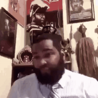 A Man With A Beard Is Sitting In A Room With A Lot Of Paintings On The Wall . GIF