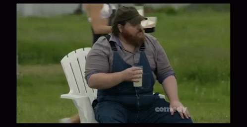 A Man With A Beard Is Sitting In A White Chair Holding A Glass Of Milk GIF