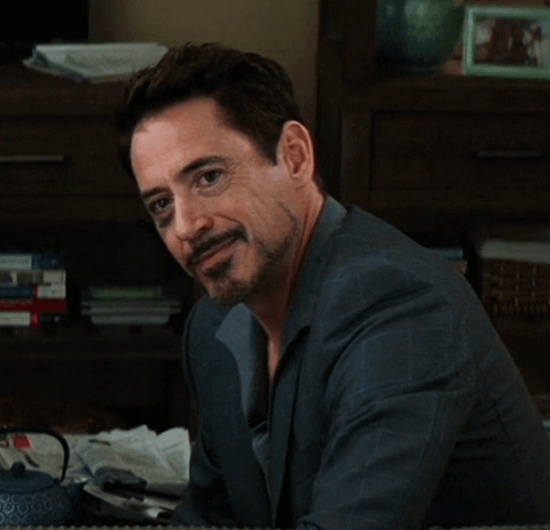 A Man With A Beard Is Sitting In Front Of A Bookshelf And Looking At The Camera GIF