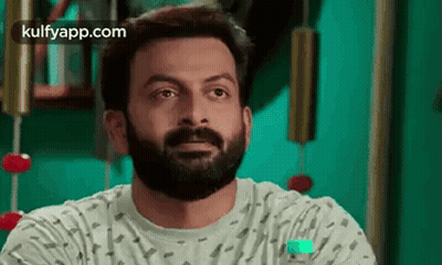 A Man With A Beard Is Sitting In Front Of A Green Wall And Looking At The Camera . GIF