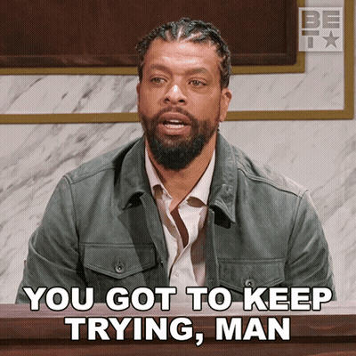 A Man With A Beard Is Sitting In Front Of A Sign That Says " You Got To Keep Trying Man " GIF