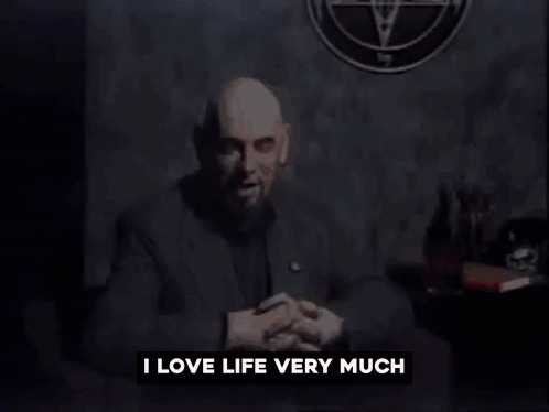 A Man With A Beard Is Sitting In Front Of A Wall That Says I Love Life Very Much GIF