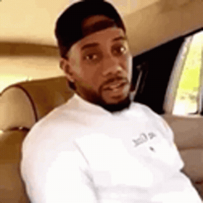 A Man With A Beard Is Sitting In The Back Seat Of A Car Wearing A Hat And A White Shirt . GIF