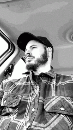 A Man With A Beard Is Sitting In The Back Seat Of A Car Wearing A Plaid Shirt And A Hat . GIF