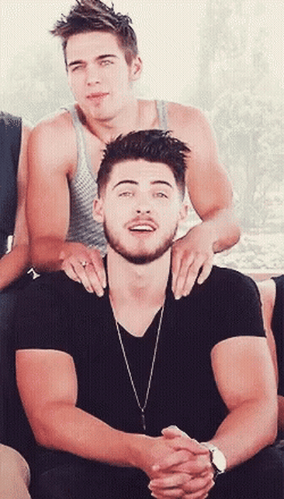 A Man With A Beard Is Sitting Next To Another Man With His Hands On His Shoulders GIF