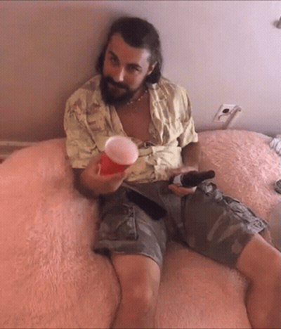 A Man With A Beard Is Sitting On A Bed Holding A Beer And A Cup GIF