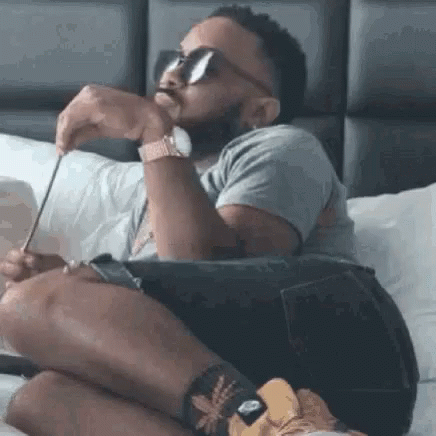 A Man With A Beard Is Sitting On A Bed Wearing Sunglasses And A Watch . GIF