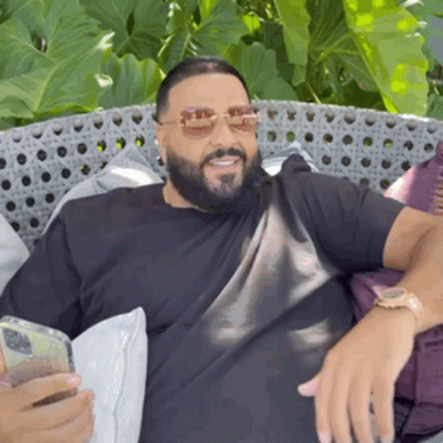 A Man With A Beard Is Sitting On A Couch Holding A Cell Phone And Wearing Sunglasses . GIF