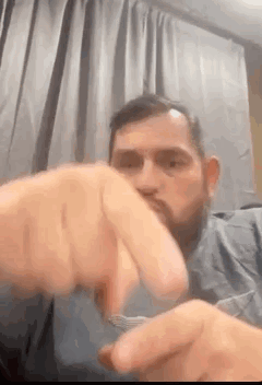 A Man With A Beard Is Sitting On A Couch Holding A Spoon In His Mouth . GIF