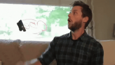 A Man With A Beard Is Sitting On A Couch In Front Of A Window Holding A Remote Control In His Hand . GIF