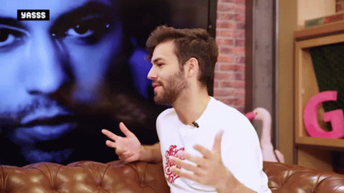 A Man With A Beard Is Sitting On A Couch In Front Of A Yasss Logo GIF