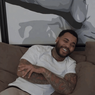 A Man With A Beard Is Sitting On A Couch Smiling GIF