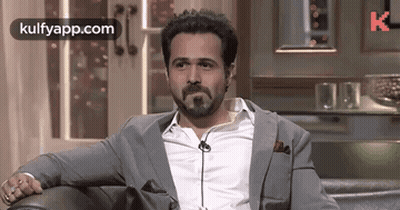 A Man With A Beard Is Sitting On A Couch Wearing A Suit And Tie . GIF