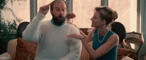 A Man With A Beard Is Sitting On A Couch While A Woman Brushes His Hair . GIF