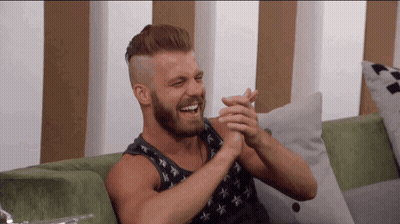 A Man With A Beard Is Sitting On A Green Couch And Clapping His Hands GIF
