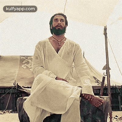 A Man With A Beard Is Sitting On A Rock In Front Of A Tent On The Beach . GIF