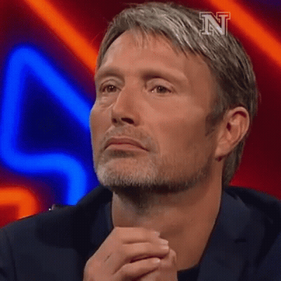 A Man With A Beard Is Sitting On A Stage With His Hands Folded In Front Of His Face . GIF