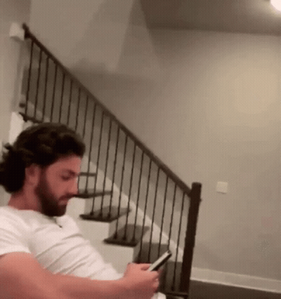 A Man With A Beard Is Sitting On A Staircase Looking At His Phone . GIF