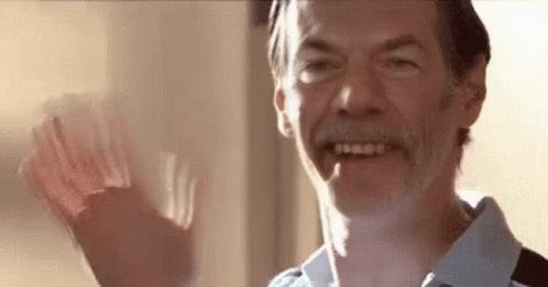 A Man With A Beard Is Smiling And Waving His Hand In Front Of A Window . GIF