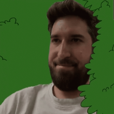 A Man With A Beard Is Smiling Behind A Green Bush GIF