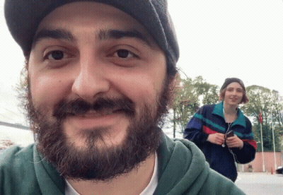 A Man With A Beard Is Smiling While A Woman Stands Behind Him Holding A Cell Phone GIF