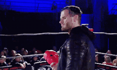 A Man With A Beard Is Speaking Into A Microphone In Front Of A Crowd Of People GIF