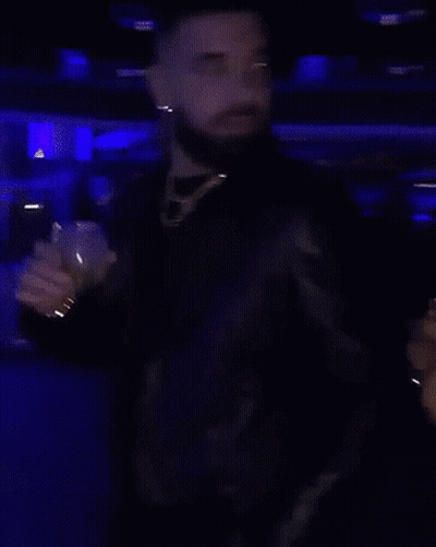 A Man With A Beard Is Standing In A Dark Room Holding A Glass GIF