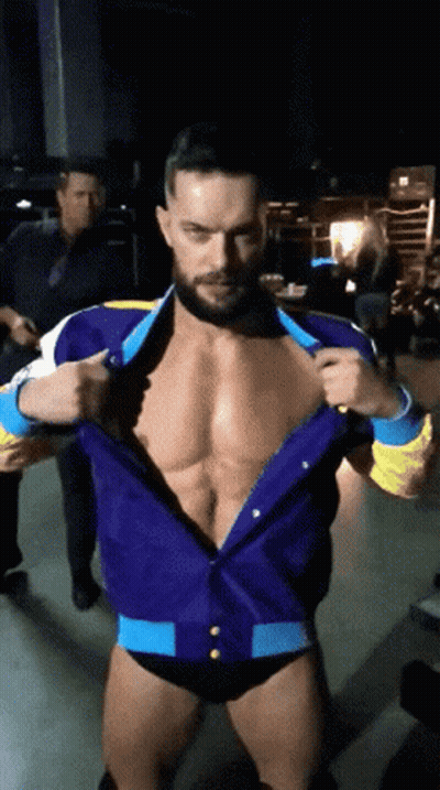 A Man With A Beard Is Standing In A Dark Room Taking Off His Shirt GIF