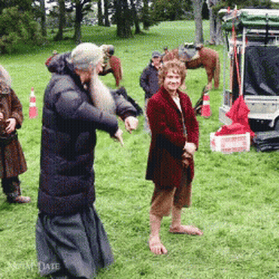 A Man With A Beard Is Standing In A Field With Other People And A Horse In The Background GIF