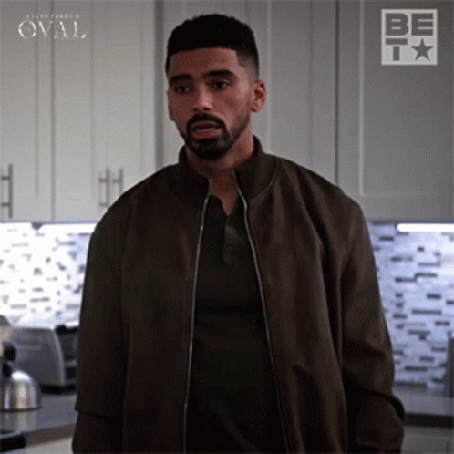 A Man With A Beard Is Standing In A Kitchen Wearing A Brown Jacket And A Black Shirt . GIF
