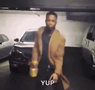 A Man With A Beard Is Standing In A Parking Garage Holding A Bottle Of Beer . GIF