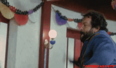 A Man With A Beard Is Standing In A Room With A Lamp And Decorations On The Wall . GIF