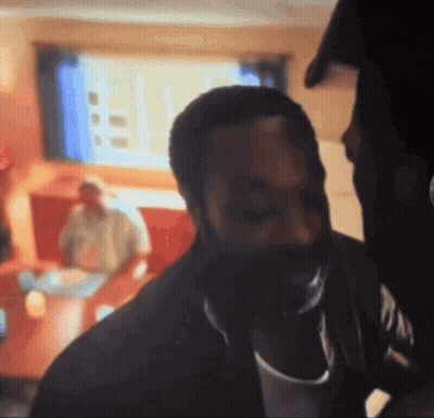 A Man With A Beard Is Standing In A Room With A Man Sitting On A Couch Behind Him . GIF
