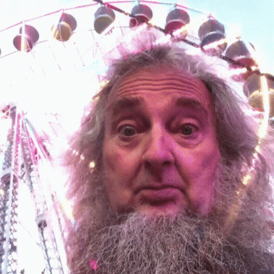A Man With A Beard Is Standing In Front Of A Colorful Ferris Wheel GIF