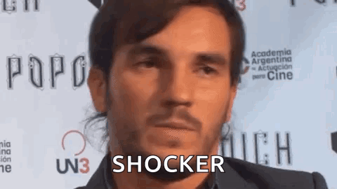 A Man With A Beard Is Standing In Front Of A Red Carpet . GIF