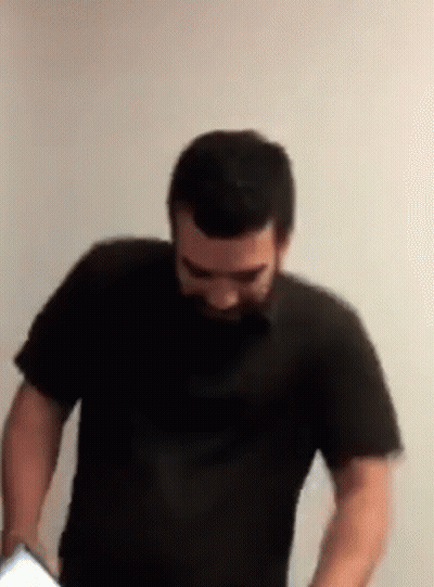 A Man With A Beard Is Standing In Front Of A White Wall Wearing A Black Shirt GIF