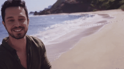 A Man With A Beard Is Standing On A Beach And Smiling GIF