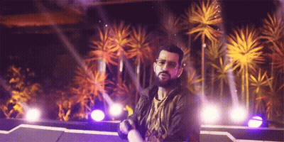 A Man With A Beard Is Standing On A Stage In Front Of Palm Trees And Purple Lights GIF