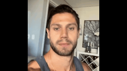 a man with a beard is taking a selfie in a living room with a wine rack in the background .