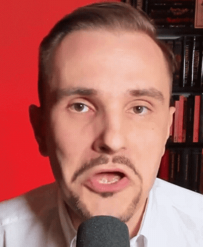 A Man With A Beard Is Talking Into A Microphone In Front Of A Bookshelf Filled With Books . GIF
