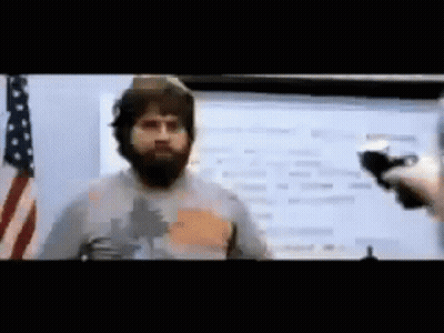 A Man With A Beard Is Talking Into A Microphone In Front Of An American Flag .. GIF