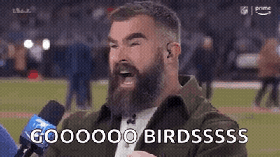A Man With A Beard Is Talking Into A Microphone On A Sports Field . GIF