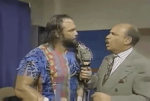 A Man With A Beard Is Talking Into A Microphone While Another Man In A Suit Stands Behind Him . GIF